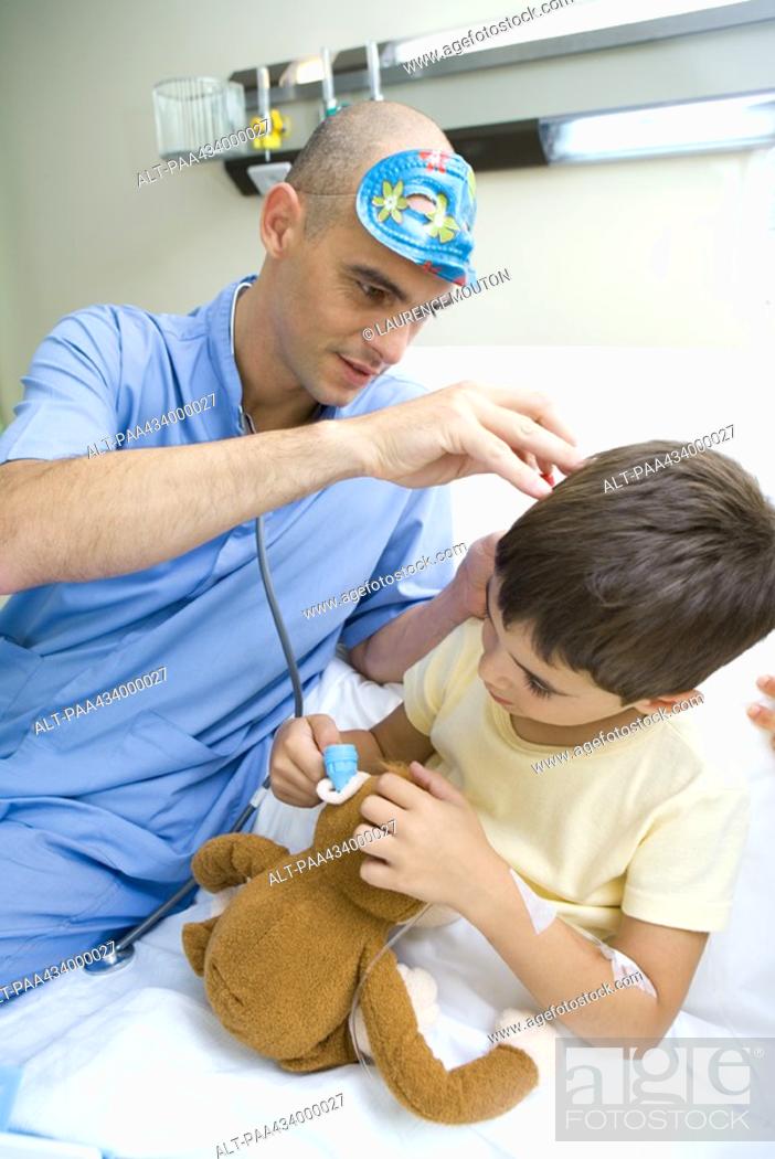 Stock Photo: Intern sitting next to boy in hospital bed, looking into boy's ear while boy imitates him with stuffed animal.