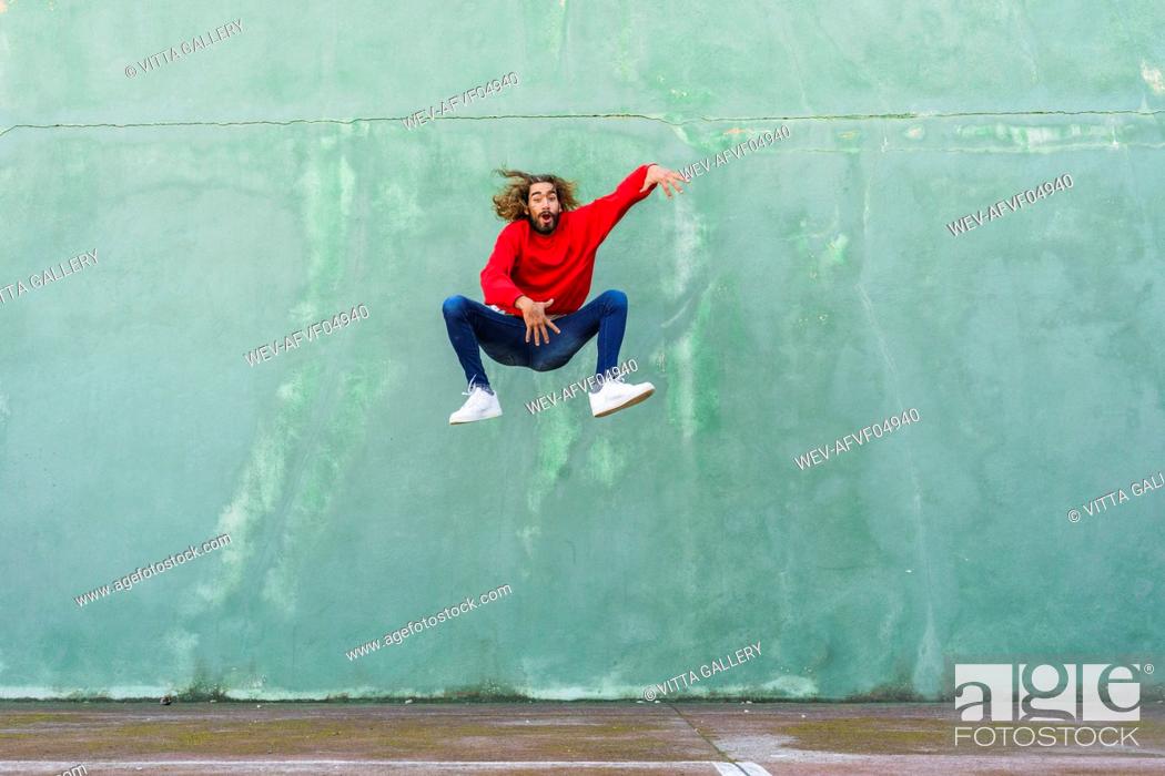 Stock Photo: Portrait of young man in wearing red sweatshirt jumping in the air in front of green wall.