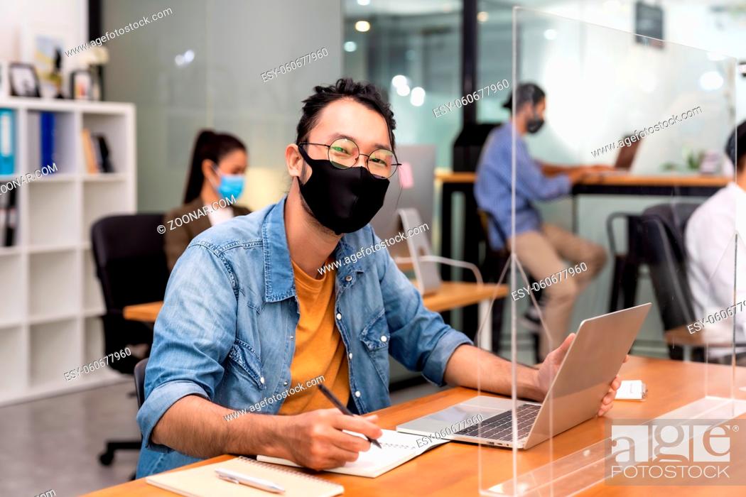 Stock Photo: Portrait of asian office employee businessman wear protective face mask work in new normal office with interracial colleague in background as social distance.