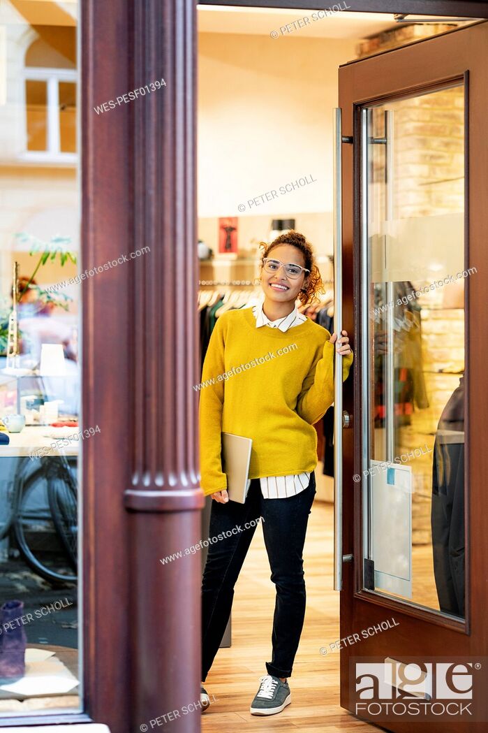 Stock Photo: Young woman standing in door of a fashion store, holding laptop.