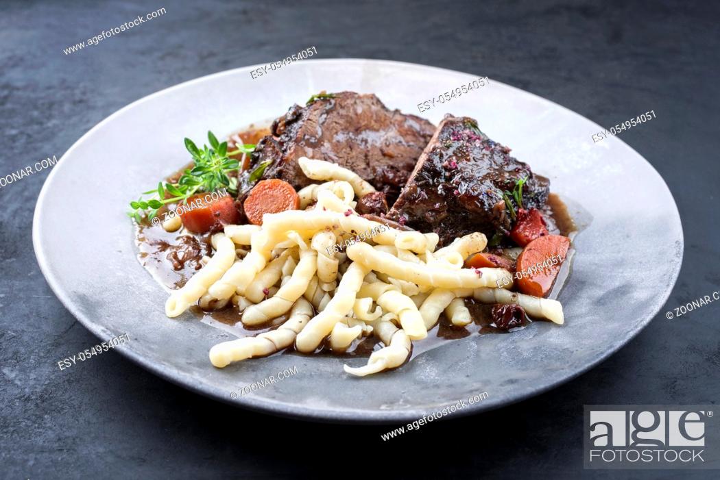 Stock Photo: Traditional German braised beef cheeks in brown red wine sauce with noodles and broccoli offered as closeup on a modern design plate on an old rustic board.