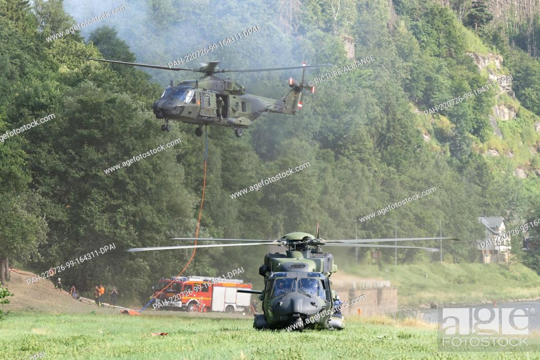 Stock Photo: 26 July 2022, Saxony, Schmilka: A German Armed Forces helicopter takes off with a firefighting water external load container on the banks of the Elbe River in.