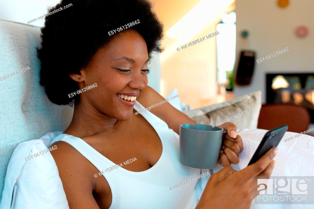 Stock Photo: Cheerful afro african american young woman having coffee while using smart phone in bedroom.