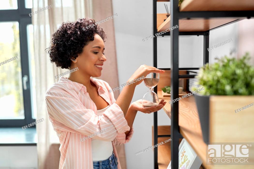 Stock Photo: smiling woman with hourglass at shelf at home.