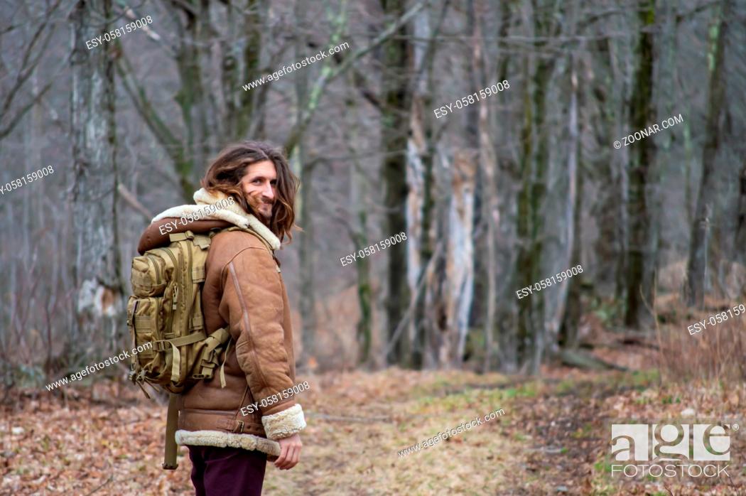 Stock Photo: Rear view of a young man with long hair, sheepskin winter coat and green military backpack looking at camera while walking in the woodlands.