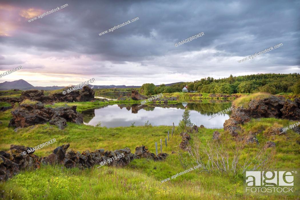 Stock Photo: Lava formations of volcanic rock rise from green meadows with dramatic skies at Kalfaströnd, house is reflected in lake Mývatn, Skútustaðir, Norðurland eystra.