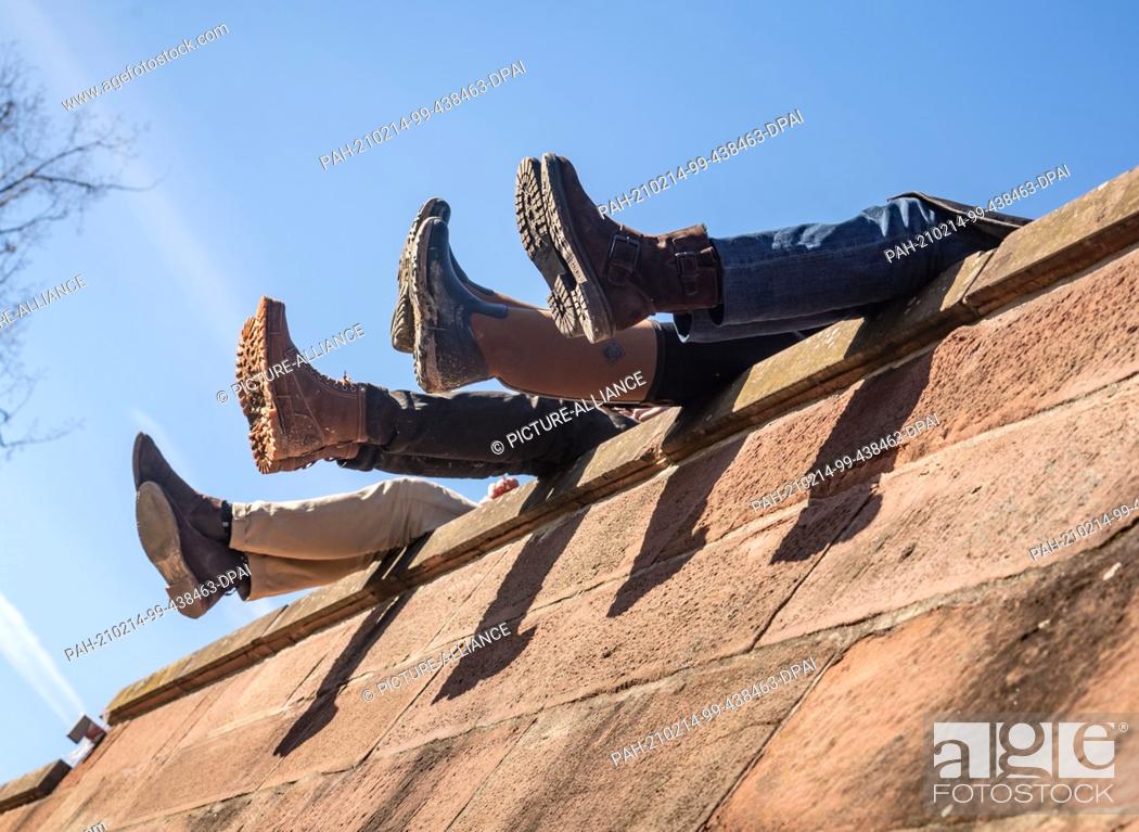 Stock Photo: 14 February 2021, Hessen, Frankfurt/Main: Four people dangle their legs from the high wall on the Mainkai in the afternoon as they sit together in the sunshine.