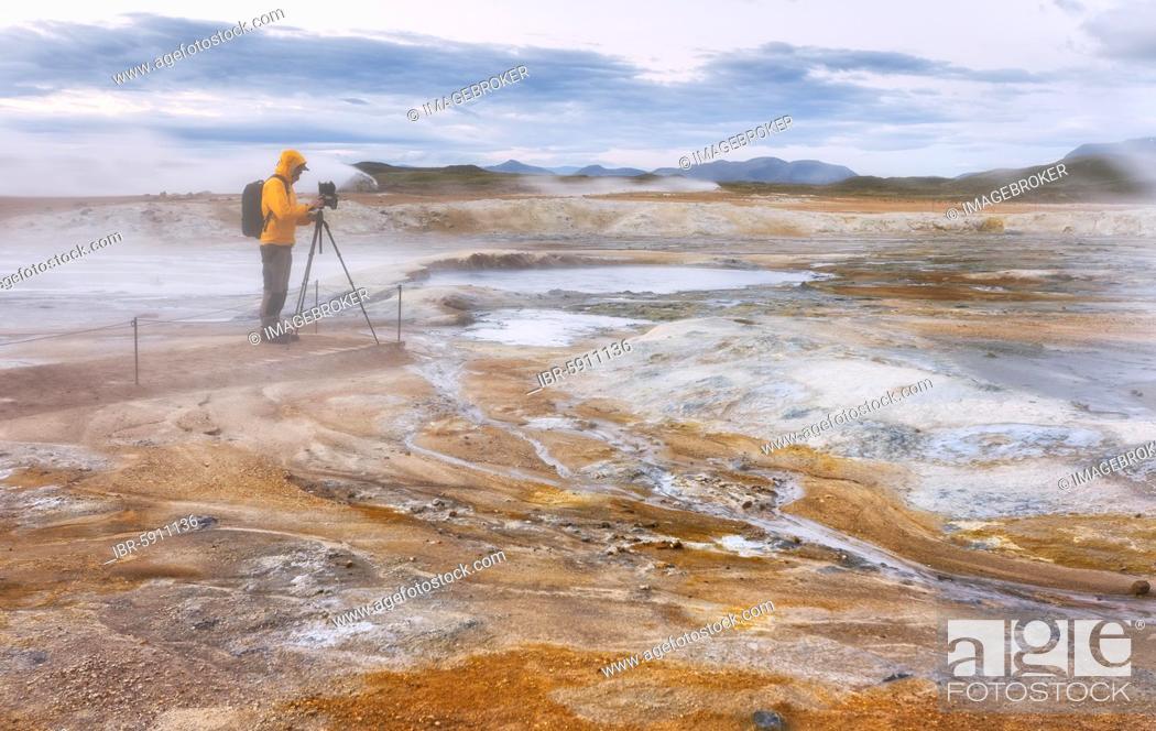 Stock Photo: Man in yellow jacket with photo tripod, photo backpack and camera standing in the steaming red high temperature area Hverarönd with solfatars and fumaroles.