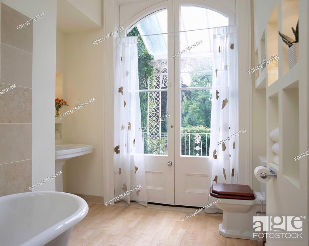 Stock Photo: Patterned white voile curtains on French doors in modern white bathroom with wooden flooring.