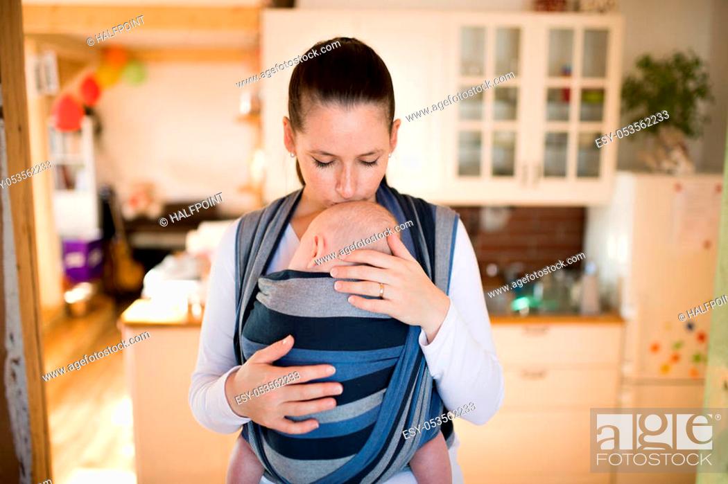 Stock Photo: Beautiful young mother in kitchen with her baby son sleeping in sling at home, kissing him on head.