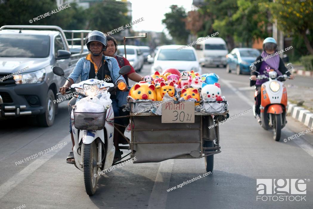 Stock Photo: 19 October 2019, Thailand, Chiang Mai: A street hawker sits on a motorcycle next to his goods and waits at a traffic light in traffic.