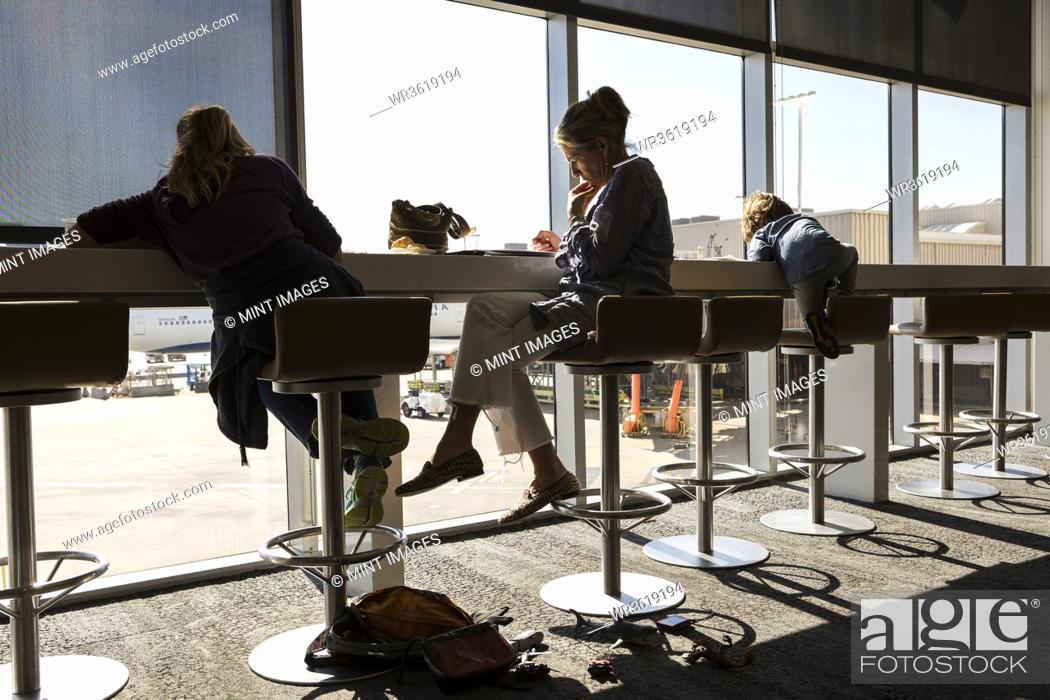 Photo de stock: A mature woman and two children seated on high stools at an airport departure lounge iwth a view over the airport apron.