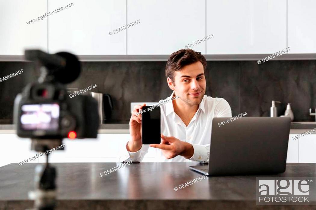 Stock Photo: Concentrated young man filming his video blog episode about new tech devices while sitting at the kitchen table and showing blank screen of mobile phone.