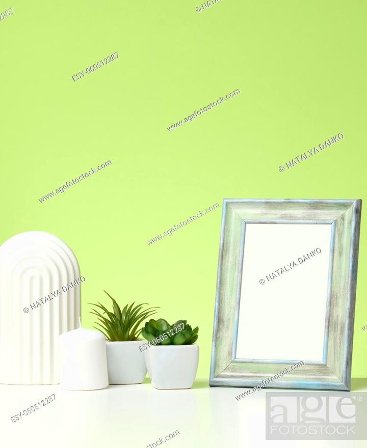 Imagen: empty white wooden photo frame and flowerpots with plants on white table, green background.