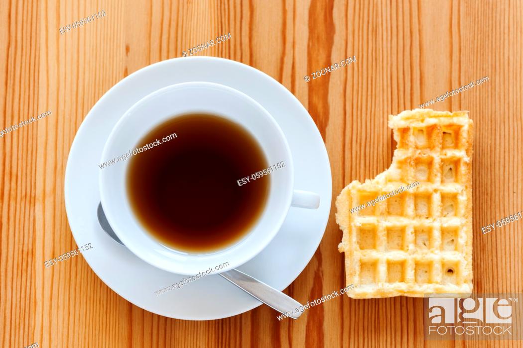 Stock Photo: Wooden table with white cup with saucer and spoon as well as lying waffle with bite mark in top view.