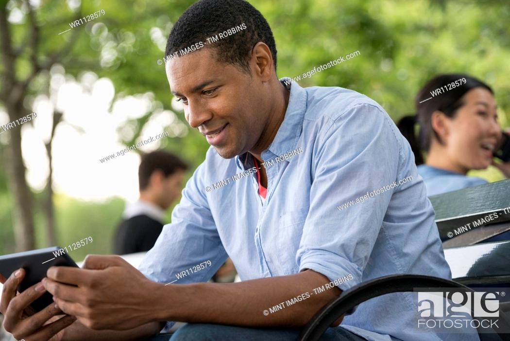 Stock Photo: Summer in the city. People outdoors, keeping in touch while on the move. A man sitting on a bench using a digital tablet.