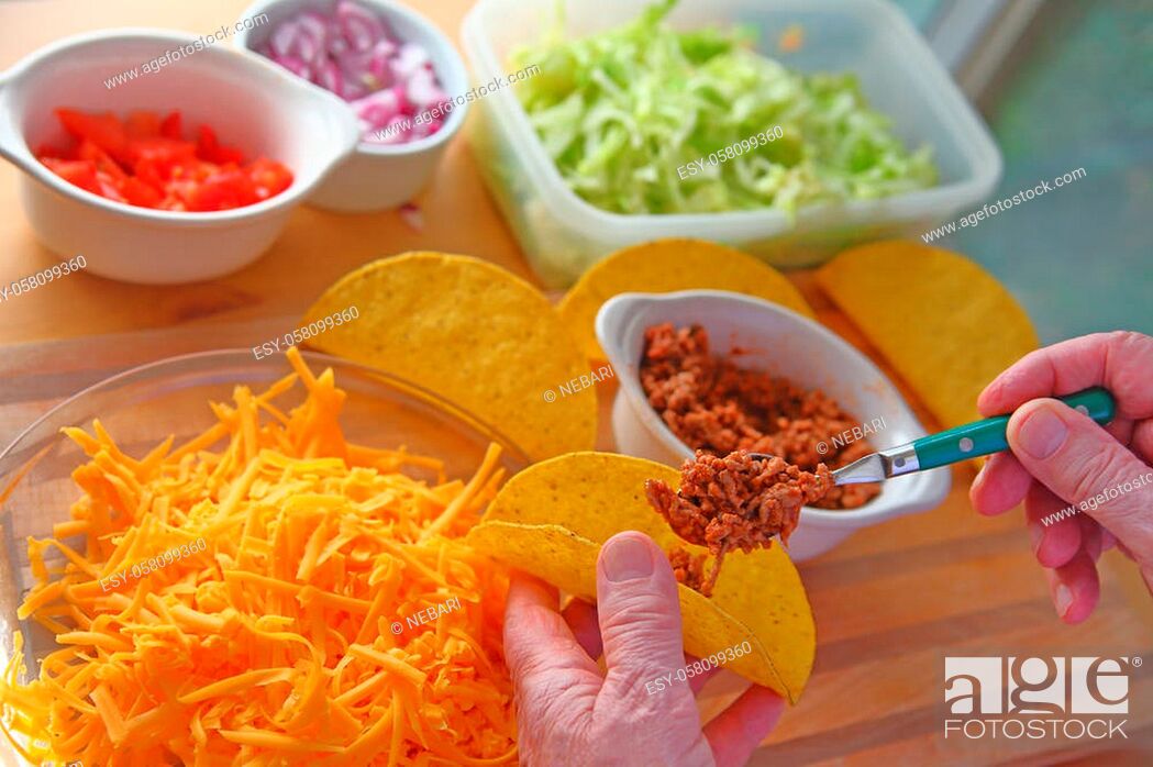 Stock Photo: A man makes tacos at home with taco shells, ground beef, lettuce, onions, tomatoes and grated cheese.