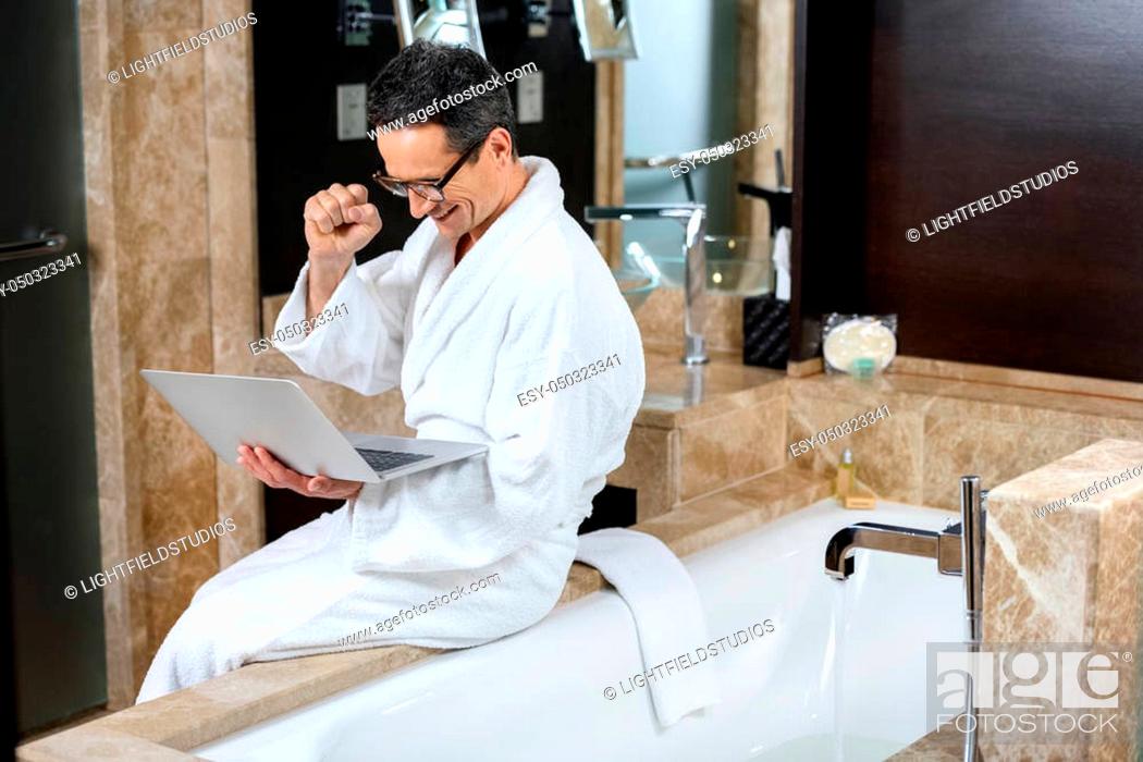 Stock Photo: Businessman wearing a bathrobe in hotel bathroom, using his laptop while sitting on bathtub.