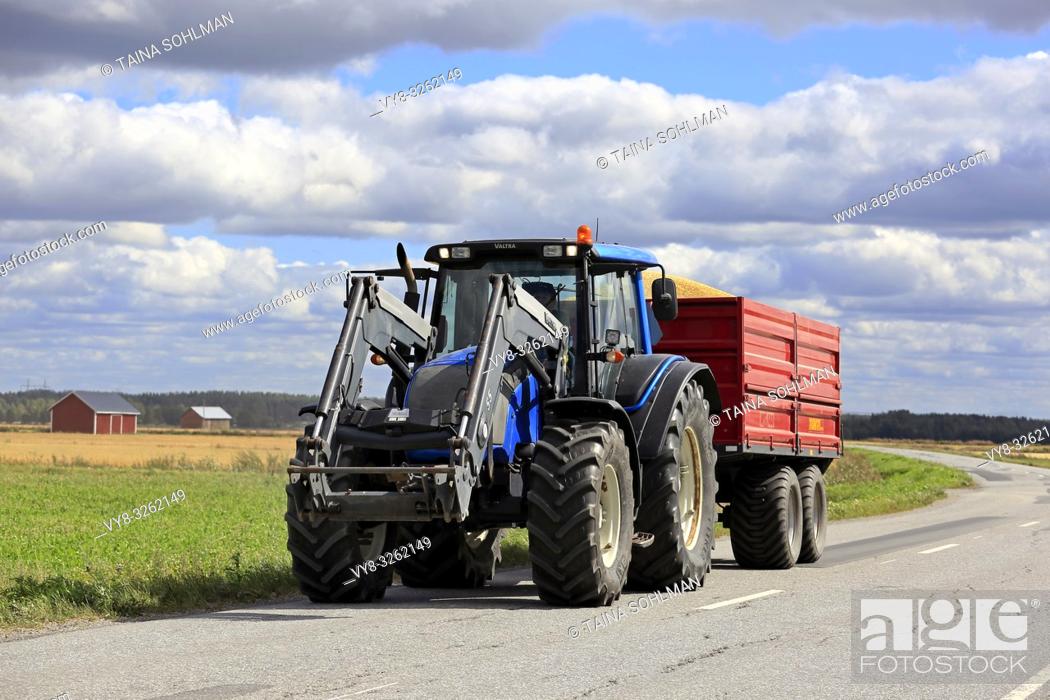 Stock Photo: Ilmajoki, Finland - August 11, 2018: Blue Valtra farm tractor pulls trailer load of harvested grain along rural road on a clear day of autumn harvest.