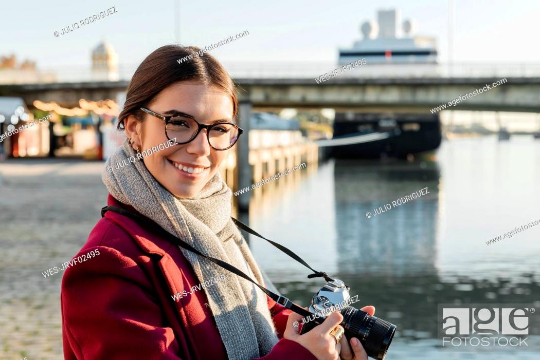 Stock Photo: Smiling young woman with camera at riverbank on sunny day.