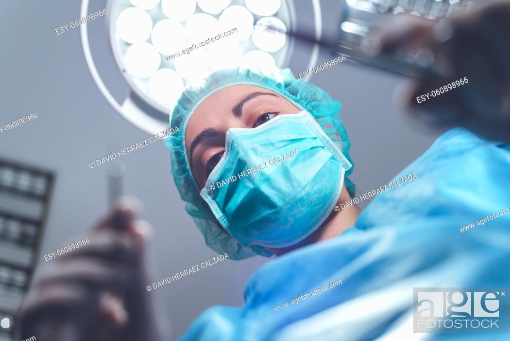 Stock Photo: From below woman surgeon in medical uniform using professional tools while standing under bright light in operating theater. High quality photo.