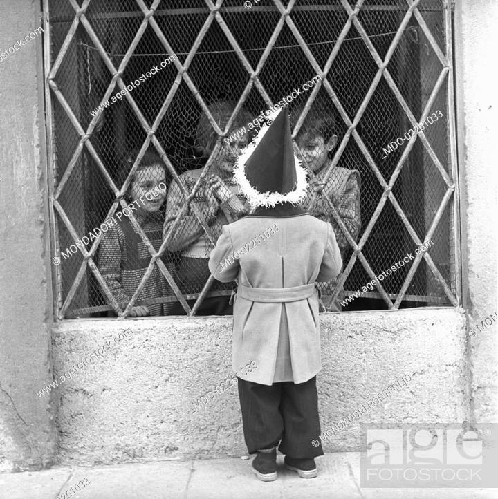 Stock Photo: A child wearing a carnival hat talking with some children behind a grating. Bergamo, 1950s.
