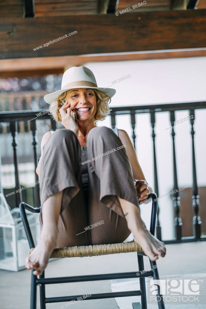 Stock Photo: Portrait of a mature caucasian blonde young woman in her 50s talking on a mobile phone smiling and wearing a hat and sitting in a relaxed pose in a rocking.