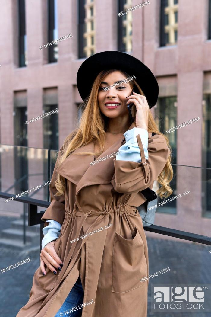 Stock Photo: Smiling woman with trenchcoat talking on mobile phone in front of glass railing.