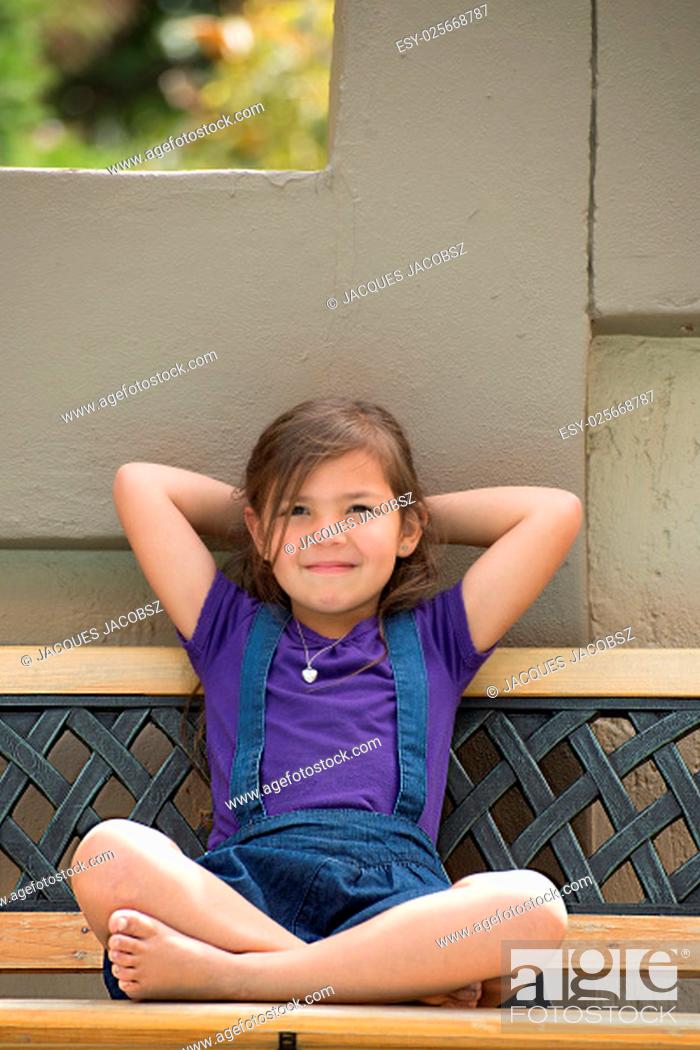 Stock Photo: A young girl sits on an outside bench with heer legs crossed, her arms behind her head and her head against the wall, while giving a slight smile.