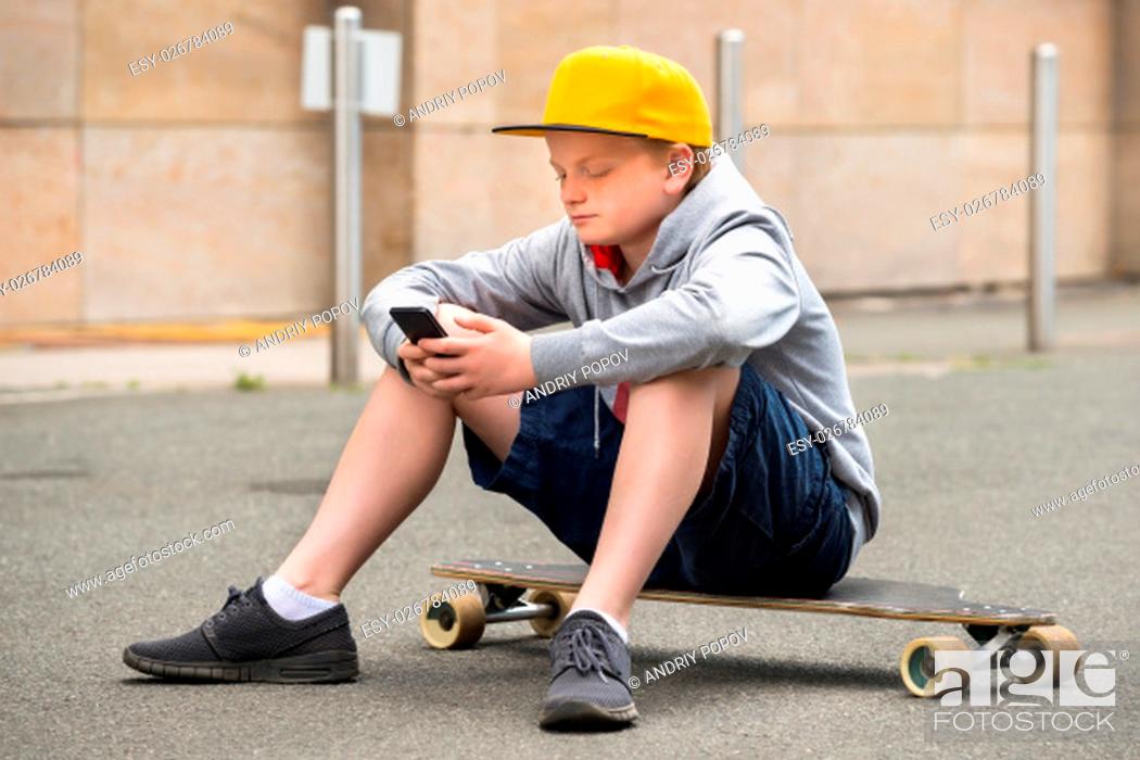 Stock Photo: Boy Wearing Cap Sitting On His Skateboard Using Smartphone.