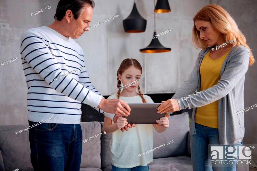 Stock Photo: Dragging from tablet. Resolute confident mature parents standing at home and taking away laptop from child while expressing discontent and upbringing child.