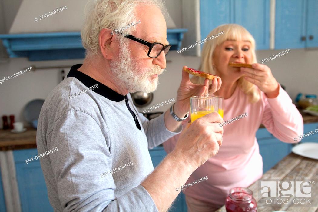 Stock Photo: Old grandpa chewing on some jam toasts with his wife at breakfast. Cute old man with glasses and white beard having some bread with jelly and orange juice for.
