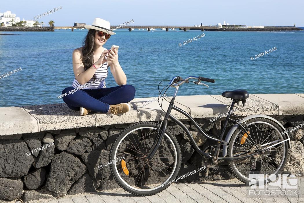 Stock Photo: Young smiley woman in hat and sunglasses using device while sitting on quay in Lanzarote, Gran Canaria.