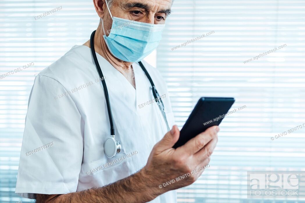 Stock Photo: Mature male doctor - nurse wearing face mask, looking at mobile phone next to a hospital window. Covid-19 and medicine concept.