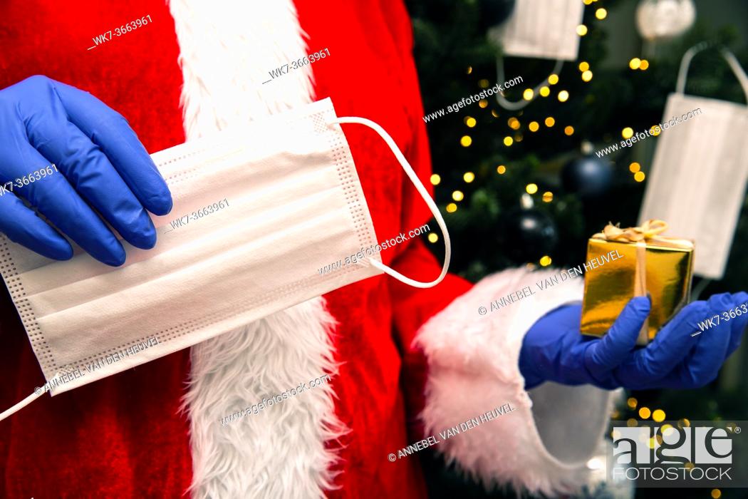 Stock Photo: Santa Claus with gloves and face mask for Coronavirus by the Christmas tree, holding a golden present, Covid-19 and Christmas safety concept closeup.