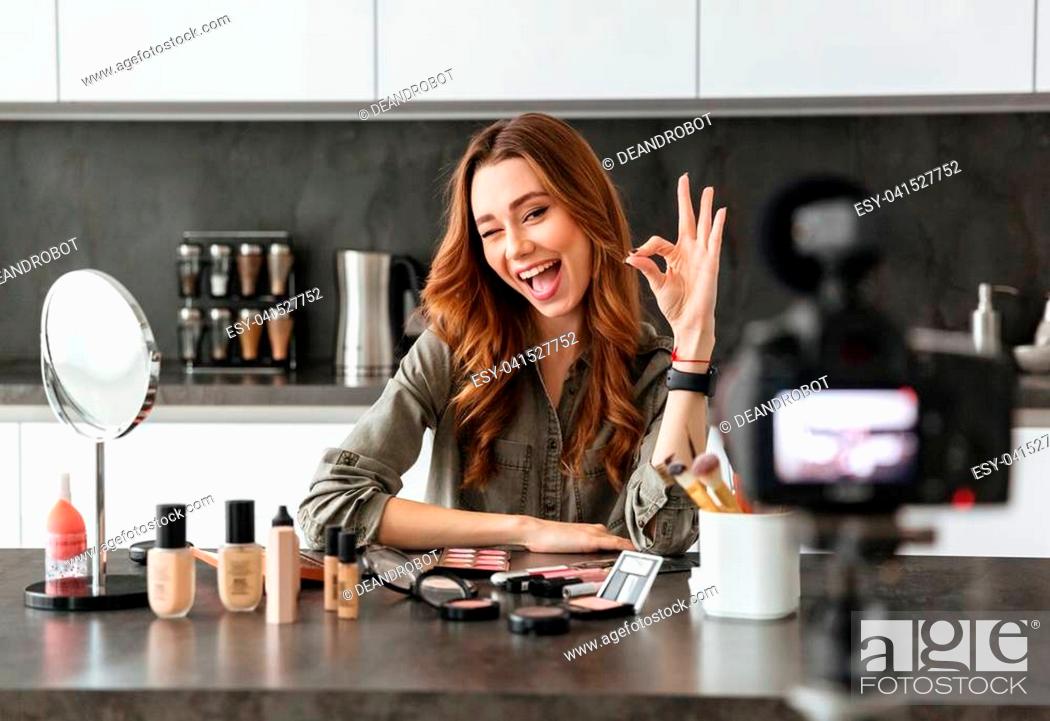 Stock Photo: Smiling young girl recording her video blog episode about new cosmetic products while sitting at the kitchen table at home and applying make-up and showing ok.