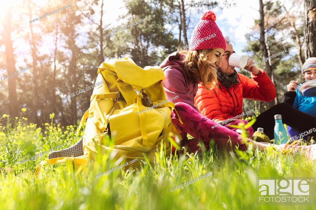 Stock Photo: Happy woman with backpack sitting by friends eating and drinking in forest on sunny day.