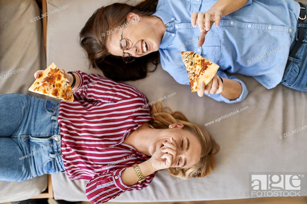 Imagen: Two laughing young women lying down eating pizza together.