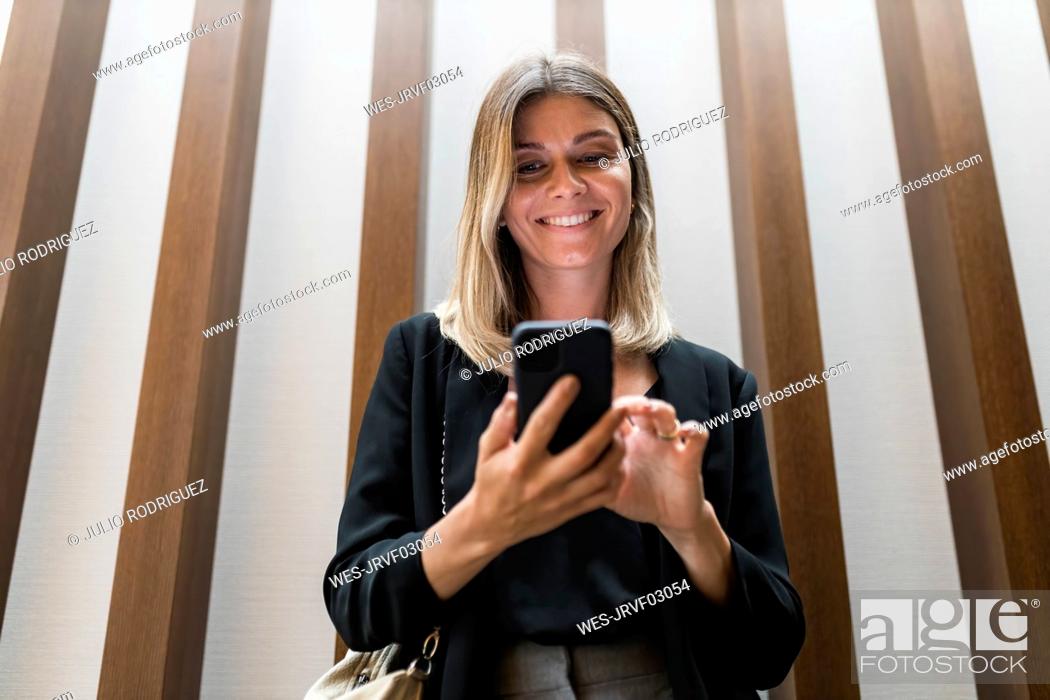 Imagen: Smiling businesswoman using smart phone in front of wall.