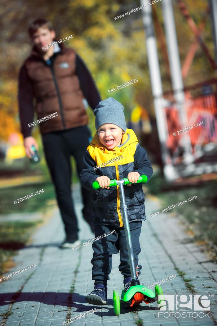 Stock Photo: A happy little boy on a scooter walks in the park with his father on an autumn day. The child rides a scooter on the sidewalk and the parent walks behind and.