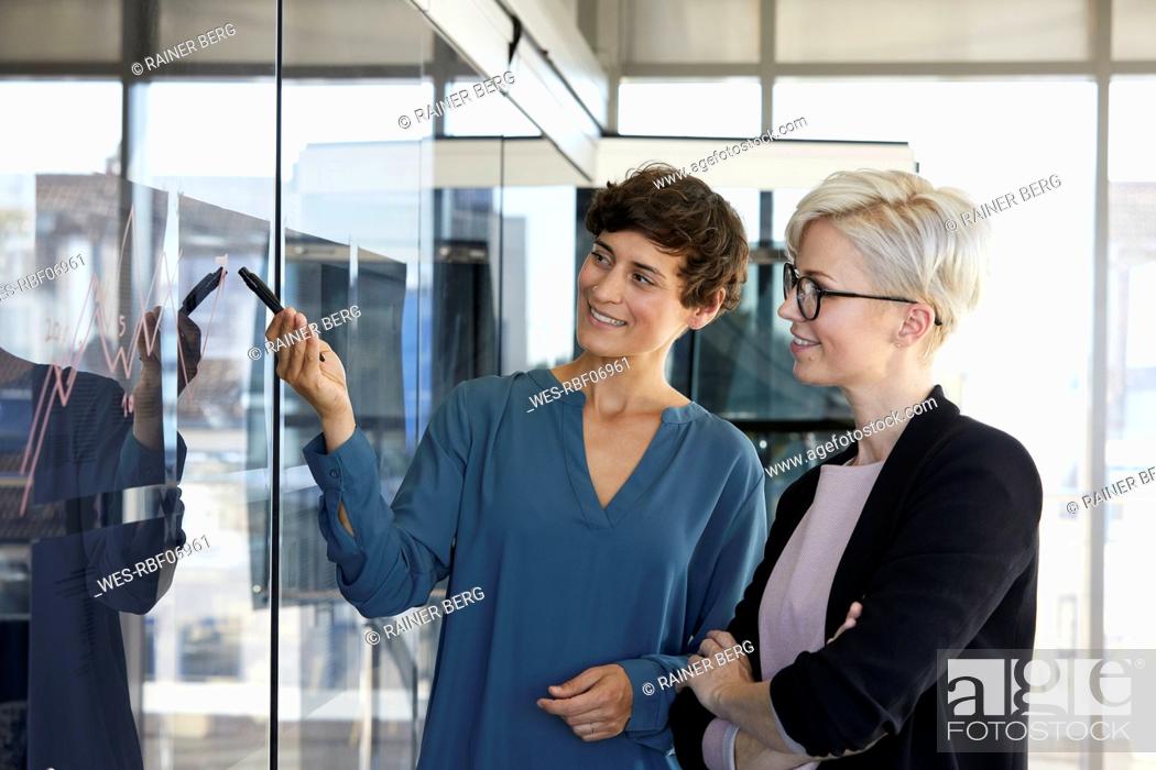Stock Photo: Two smiling businesswomen looking at chart on glass pane in office.