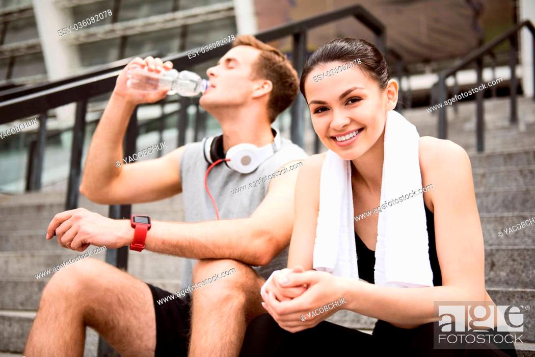 Stock Photo: Get some relaxation. Cute young woman looking at camera near a man drinking water while having a rest after exercising.