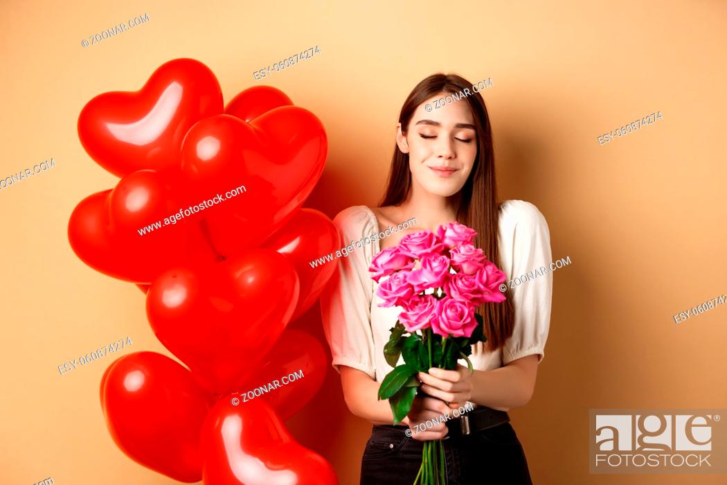 Stock Photo: Romantic girlfriend smelling roses, receive flowers from lover on Valentines day, standing near red hearts balloons and smililng, beige background.