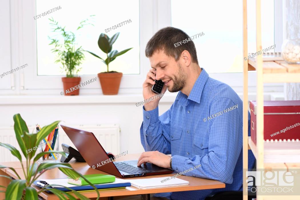Stock Photo: Young businessman talking on the phone in the office and working in a laptop.