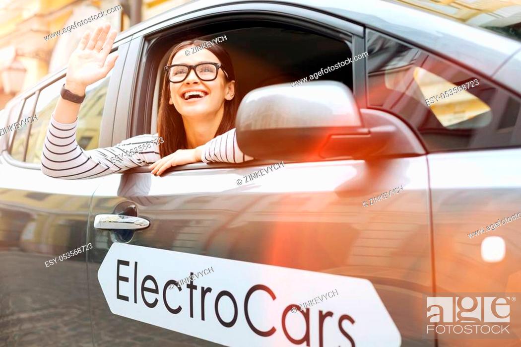 Stock Photo: Happy to see you. Gorgeous young woman in glasses sitting in the electric car, looking out of a window and waving at someone familiar in the street.
