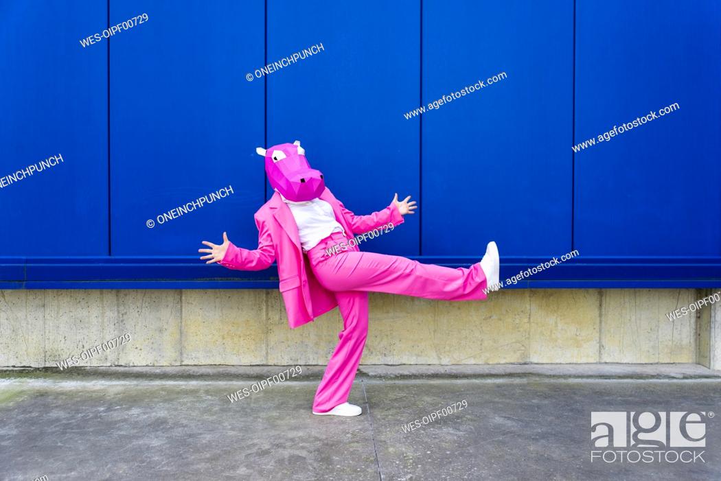 Stock Photo: Woman wearing vibrant pink suit and hippo mask posing on one leg in front of blue wall.