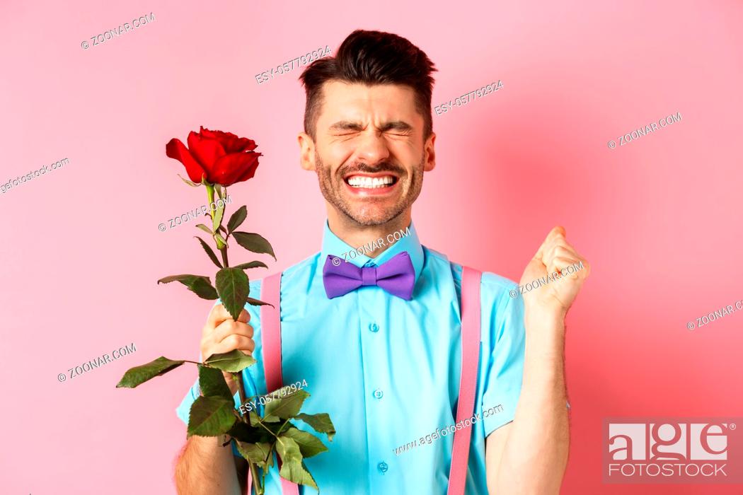Imagen: Romance and Valentines day concept. Hopeful man feeling excited before date, waiting for lover with red rose and praying, standing over pink background.
