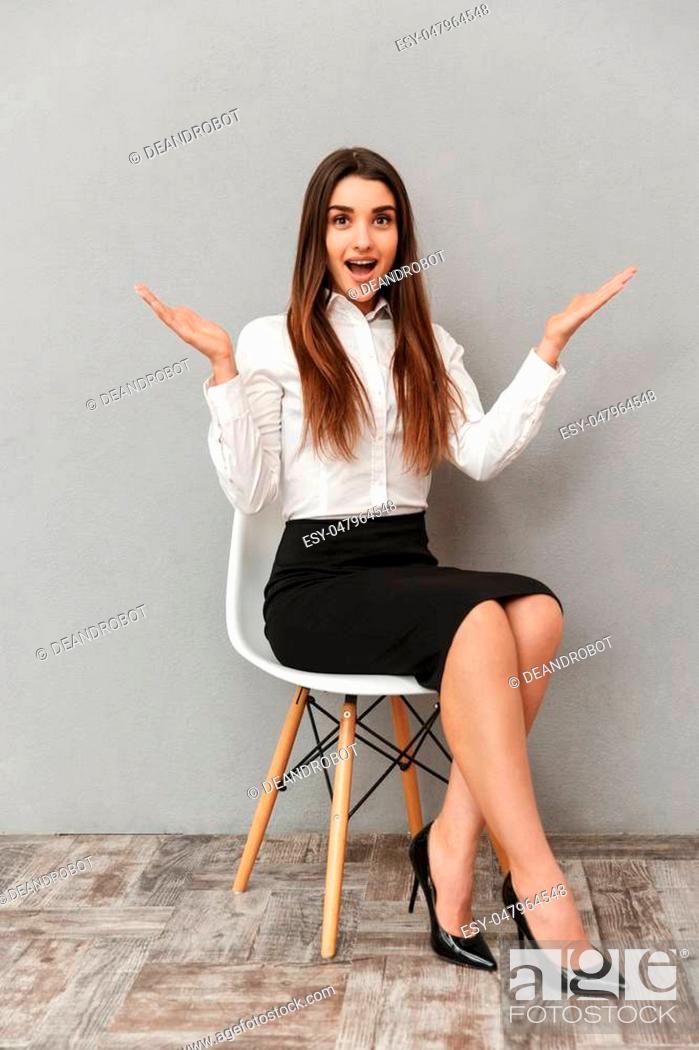 Stock Photo: Full length portrait of joyful pretty woman with long brown hair in business wear throwing up hands while sitting on chair isolated over gray background.