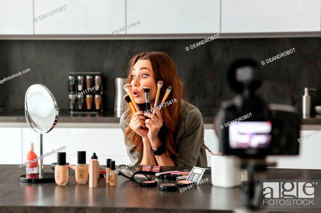 Stock Photo: Satisfied young girl recording her video blog episode about new cosmetic products while sitting at the kitchen table at home and applying make-up.