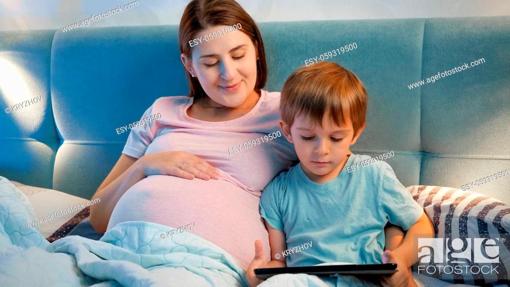 Stock Photo: Portrait of pregnant young mother hugging her little son lying in bed and playing on tablet computer before going to sleep.
